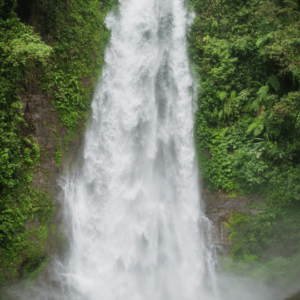 A close-up of a powerful waterfall cascading down a rocky cliff face, surrounded by lush green vegetation. The water creates a mist as it falls