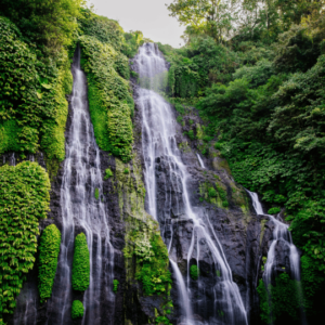 A breathtaking view of twin waterfalls cascading down a lush, green cliff face. The water appears milky white as it plunges into a series of rock pools below