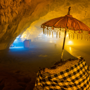 Dimly lit interior of Goa Giri Putri cave in Bali, featuring a traditional Balinese umbrella shading a small shrine wrapped in a checkered cloth, illuminated by soft, warm lighting