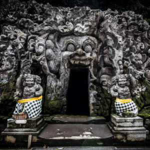 Entrance to Goa Gajah, the Elephant Cave in Bali, featuring an intricately carved stone face and two statues dressed in traditional Balinese sarongs.