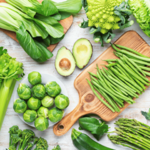 An assortment of fresh green vegetables arranged on a light wooden surface. The selection includes bok choy, avocado halves, Brussels sprouts, celery, green beans, zucchini, asparagus, romanesco broccoli, and leafy greens. The vegetables are displayed on wooden cutting boards and in bowls, highlighting their vibrant colors and fresh textures