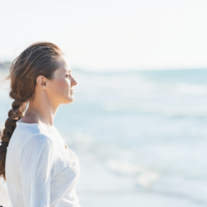 A serene image of a woman standing by the ocean, looking out at the water. She has her hair tied back in a braid and is wearing a light, long-sleeved top. The soft, natural light and blurred background of the sea create a peaceful and reflective atmosphere