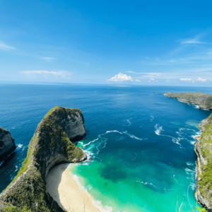 A breathtaking view of a coastal cliff formation at Kelingking Beach on Nusa Penida, Bali. The turquoise waters of the ocean gently meet the sandy beach below, framed by steep cliffs covered in green vegetation. The sky is clear and blue, with a few scattered clouds on the horizon. This image highlights the natural beauty of Bali, a popular destination accessible by fast boat