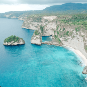 Aerial view of Diamond Beach in Nusa Penida, Bali, Indonesia. The beach is surrounded by white cliffs and turquoise blue water. There is a small island in the middle of the bay. The sky is cloudy