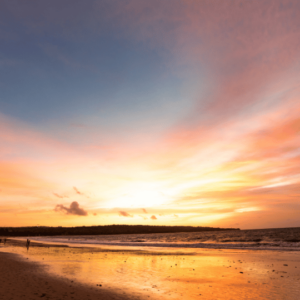A breathtaking sunset over Jimbaran Beach in Bali. The sky is ablaze with vibrant hues of orange, pink, and purple, reflecting on the calm ocean water. Silhouettes of people can be seen strolling along the beach in the foreground