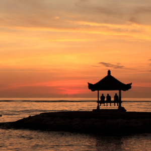 A group of people are silhouetted against a vibrant orange sunset as they sit in a traditional Balinese gazebo on the beach. The ocean is calm and reflects the warm colors of the sky